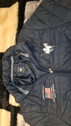 Mörkblå quiltad jacka Vinson Polo Club - Quiltad mörkblå pufferjacka från Vinson Polo Club med broderad logga och amerikansk flagga på bröstet. Jackan har dragkedja framtill, två sidofickor med dragkedja och ståkrage. Perfekt för kyligare dagar.