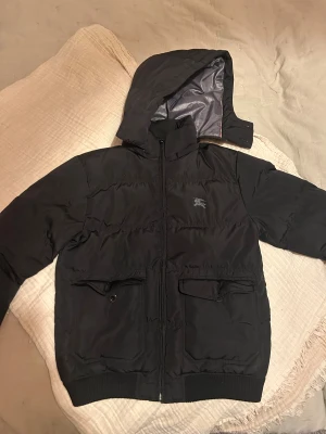 Svart pufferjacka med huva Burberry - Svart pufferjacka från Burberry med huva och dragkedja framtill. Jackan har två stora fickor med lock och knapp, ribbade muddar och diskret Burberry-logga på bröstet. Perfekt för kalla dagar och riktigt snygg till streetwear.