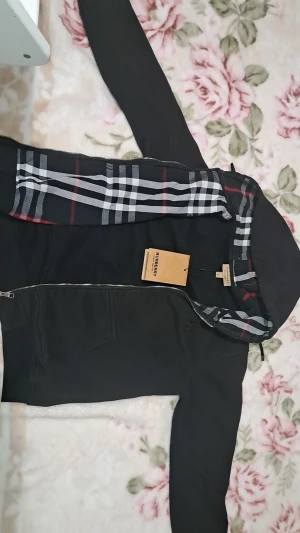 Svart Burberry hoodie med rutig huva - Svart hoodiejacka från Burberry med dragkedja, känguruficka och klassiskt rutig huva i svart, vitt och rött. Jackan har snörning i huvan och är tillverkad i mjuk bomull med fleecefoder. Perfekt för dig som vill ha en stilren och ikonisk look. Helt ny etikett på
