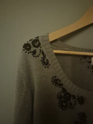 Grå kofta med blommigt mönster från Rabalder - Snygg grå kofta från Rabalder i storlek M. Den har svarta blommiga detaljer på axlar, bröst och nederkant, rund halsringning och knappar framtill. Materialet är stickad och känns mjukt mot huden. Perfekt för dig som gillar en chill och feminin stil! 