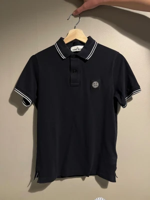 Svart pikétröja Stone Island - Svart pikétröja från Stone Island med vit kontrast på krage och ärmslut.  och ikonisk logopatch på bröstet. Tillverkad i mjuk bomull, perfekt för en clean och stilren look.
