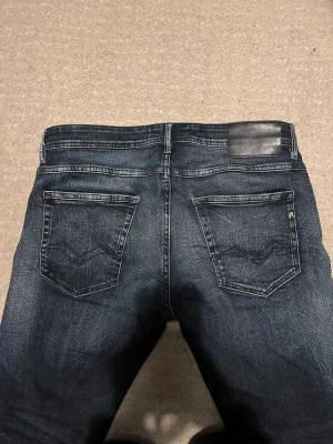 Replay Anbass mörkblå jeans - Snygga Replay Anbass jeans i mörkblå tvätt med diskreta slitningar och klassiska fem fickor. Modellen har smal passform och raka ben, med Replay-logga på fickan och svart läderpatch bak i midjan. Perfekt för dig som gillar stilrena jeans med lite edge.