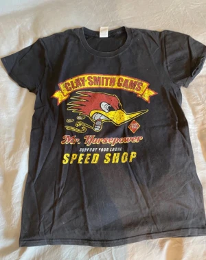 Svart t-shirt Clay Smith Cams S - Cool svart t-shirt från Fruit of the Loom med färgglatt tryck av Clay Smith Cams och Mr. Horsepower på bröstet. T-shirten har korta ärmar och rund hals. Materialet är mjuk bomull och trycket har detaljer i rött och gult som sticker ut.