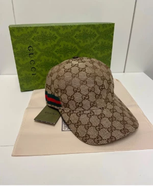 Gucci  - Snygg beige keps från Gucci med klassiskt GG-monogram över hela kepsen och en ikonisk grön-röd rand på sidan. Kepsen har böjd skärm och justerbar rem baktill. Materialet är bomull och polyester, perfekt för dig som vill ha en lyxig streetstyle-look.