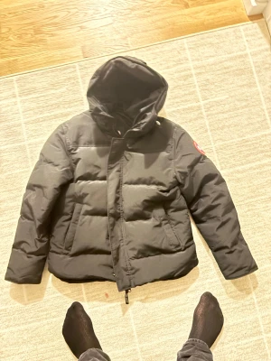 Svart pufferjacka från Canada Goose - Säljer en svart pufferjacka från Canada Goose med huva och klassisk logga på ärmen. Jackan är vadderad, har dragkedja och stora fickor framtill. Perfekt för kalla dagar och har en clean, stilren look som funkar till det mesta.