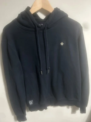 Mörkblå hoodie från Morris, strl M - Snygg mörkblå hoodie från Morris med klassisk huva och snörning. Tröjan har ett litet broderat emblem på bröstet och ribbade muddar vid ärmslut och nederkant. Perfekt för chill dagar och enkel att matcha med jeans eller joggers.