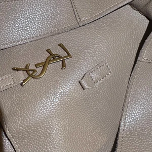 Beige axelväska från Saint Laurent - Snygg beige axelväska från Saint Laurent med klassisk YSL-logga i guld på framsidan. Väskan är tillverkad i strukturerat skinn och har en stilren design med bred axelrem och praktisk dragkedja. Perfekt för dig som gillar lyxiga detaljer och tidlös stil.