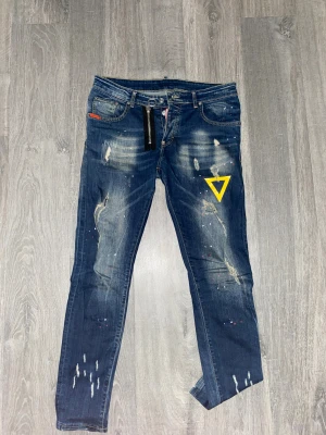 Dsquared2 jeans - Snygga blå jeans från Dsquared2. Jeansen har slitna detaljer, färgstänk och en gul tryckt symbol på ena benet. Klassisk femficksmodell med dragkedja och knapp, samt Dsquared2-logga på bakfickan. Perfekt för dig som gillar streetstyle.