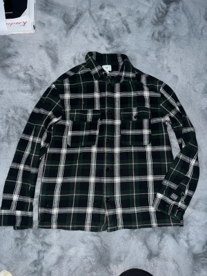 Rutig skjorta från H&M i grönt och svart - Säljer en rutig skjorta från H&M i grönt, svart och vitt med diskreta rosa linjer. Skjortan har två bröstfickor med knappar, klassisk krage och långa ärmar. Perfekt för dig som gillar chill och avslappnad stil.