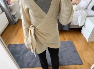 Beige tröja med knyt - Säljer en beige stickad tröja/kofta med snygg knyt i sidan och v-ringad hals. Tröjan har långa ärmar och är perfekt för lager-på-lager. 