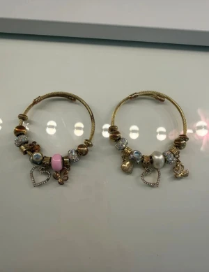 Två guldiga charmarmband med berlocker - Två guldiga armband med flera berlocker, bland annat hjärtan med strass, pärlor, glittriga kulor och detaljer i rosa och vitt. En för 15 kronor. Går att anpassa storlek