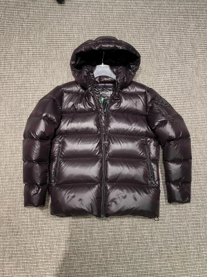 Snoot jacka (ny) - Snygg svart dunjacka från Snoot Stockholm med huva och flera praktiska fickor med dragkedja. Jackan har en glansig finish, patch på ärmen och är quiltad för extra värme. Perfekt för kalla dagar och har en cool limited edition-detalj på ärmen.LIMITED EDITION 