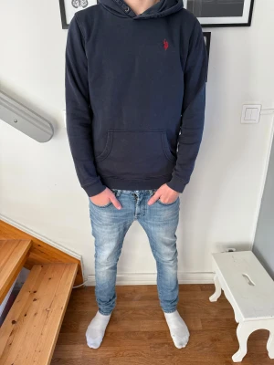Mörkblå hoodie från U.S. Polo Assn. - Snygg mörkblå hoodie från U.S. Polo Assn. med röd logga broderad på bröstet. Tröjan har huva med snörning, magficka och långa ärmar. Tillverkad i mjuk bomullsmix som är skön att ha på sig. Perfekt för chill dagar och häng med kompisar.