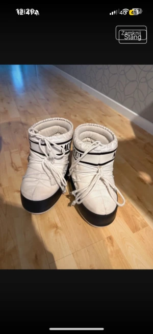 Vita Moon Boots med snörning - Moon Boots i vit färg med svart detalj och logga runt skaftet. Skorna har snörning framtill och en rund tå. Yttermaterialet är syntet och sula i gummi med mönstrad undersida för extra grepp. Perfekta för vintern och riktigt snygga till pufferjacka.