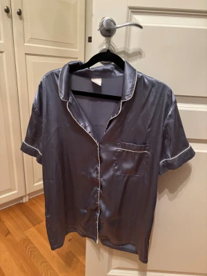 Sovtröja - Snygg blåsilvrig skjorta i glansig polyester med korta ärmar och vit piping längs kanterna. Klassisk krage och bröstficka med vit kant. Perfekt för dig som gillar en chill och stilren look.