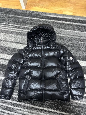Moncler jacka  - Säljer en svart Moncler Maya jacka med huva och dragkedja framtill. Jackan har två snedställda fickor med dragkedja och klassisk Moncler-logga på ärmen. NFC funkar. Passar M/L Perfekt för kalla dagar och riktigt snygg streetstil. Pris kan diskuteras vid snabb affär.