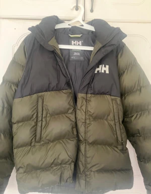 Grön och svart pufferjacka Helly Hansen - Snygg pufferjacka från Helly Hansen i olivgrönt och svart med stor huva och dragkedja. Jackan har HH-logga på bröstet och Helly Hansen-tryck på ryggen. Perfekt för kalla dagar med sin vadderade design och praktiska fickor.