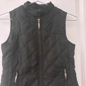 Svart quiltad ridväst Active Riding Wear - Quiltad svart väst från Active Riding Wear med broderad logga på bröstet och två fickor med dragkedja. Hög krage och hel dragkedja framtill. Perfekt för dig som gillar ridning och vill ha en stilren look.