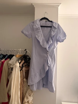 Blåvit omlottklänning med volang - Supersöt kort omlottklänning i blått och vitt med tunna ränder, volangkant längs v-ringningen och knäppning framtill. Klänningen har korta ärmar.