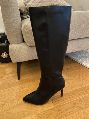 Svarta höga stövlar med klack - Säljer ett par svarta höga stövlar i skinnimitation med spetsig tå och smal hög klack. Stövlarna har dragkedja på insidan. Stövlarna är från Nelly och har utgått från deras kollektion. Jag har endast använt stövlarna vid ett tillfälle. 