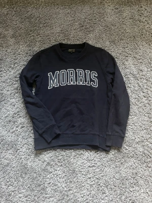 Mörkblå sweatshirt från Morris - Snygg mörkblå sweatshirt från Morris i storlek M med vit tryckt logga över bröstet. Tröjan har rund hals, ribbade muddar och är långärmad. Perfekt för dig som gillar klassisk college-stil och vill ha en bekväm tröja i mjuk bomull.
