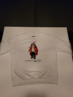 Vit sweatshirt Polo Ralph Lauren - Vit sweatshirt från Ralph Lauren med Polo Bear-tryck framtill. Björnen har röd jacka, stickad mössa och rutiga byxor. Tröjan har rund hals och långa ärmar, tillverkad i mjuk bomull. Perfekt för dig som gillar klassisk streetstyle med en twist.