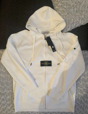 Vit hoodie från Stone Island ziphoodie - Vit hoodie från Stone Island med klassisk kompasspatch på ärmen. Tröjan har huva med snörning, ribbade muddar och hel dragkedja framtill. Tillverkad i mjuk bomull för skön komfort. Perfekt för dig som gillar clean och stilren streetwear. Helt ny för den ej passade mig riktigt😃