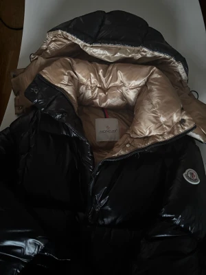 Svart pufferjacka från Moncler - Säljer en fet svart pufferjacka från Moncler med glansig finish och beige insida. Jackan har stor huva, dragkedja framtill och coola Moncler-detaljer på ärmen och dragkedjorna. Perfekt för kalla dagar när du vill se både stylish och chill ut.