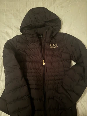 Svart pufferjacka från Emporio Armani - Svart pufferjacka från Emporio Armani EA7 med huva och guldfärgad logga på bröstet. Jackan har quiltad design, dragkedja med EA7-detalj och är perfekt för kyliga dagar. Snygg och stilren modell som håller dig varm.