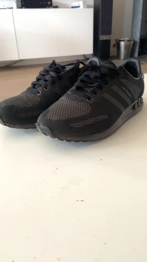 Svarta Adidas sneakers storlek 42 - Svarta Adidas sneakers med stickad ovandel och klassiska tre ränder på sidan. Skorna har snörning och platt sula med dämpande detaljer på sidan. Perfekta för dig som gillar en sportig och clean look. Märkta med 'LA TRAINER 3' på sidan.