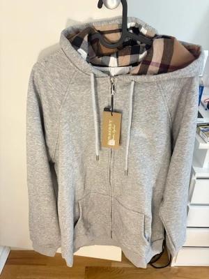 Grå hoodie från Burberry med dragkedja - Snygg grå hoodie från Burberry med klassiskt rutigt foder i huvan. Tröjan har dragkedja framtill, snörning vid huvan och två fickor. Perfekt för dig som gillar stilrena och bekväma plagg med exklusiv känsla. Storlek: M 
