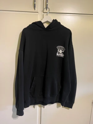 Svart hoodie från Butter med tryck - Svart oversized hoodie från Butter med stort vitt grafiskt tryck på ryggen och mindre tryck på bröstet. Tröjan har huva, känguruficka och långa ärmar. Materialet är mjuk bomull, perfekt för chill dagar. Snygg streetwear-vibe och riktigt skön passform. Köpt för ca 1 år sedan för 800kr. Se bild 4 för små fläckarna på de vita trycket på ryggen. Tveka inte på att höra av er vid funderingar