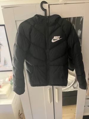 Svart pufferjacka från Nike med huva - Svart pufferjacka från Nike med stor vit logga på bröstet. Jackan har huva, dragkedja framtill och två fickor. Insidan har ett mönstrat foder i svart och vitt. Perfekt för kalla dagar och riktigt snygg streetstil. Storlek 147-158 cm eller 12-13 yrs. Köptes ny för 1200kr har används väll men inga tydliga tecken på det.