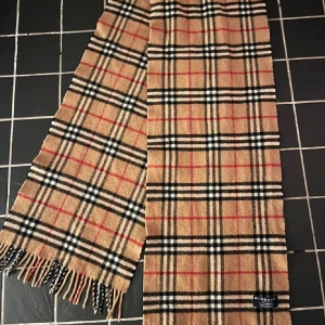 Rutig halsduk från Burberry - Säljer en klassisk rutig halsduk från Burberry i 100% lammull. Den har en snygg beige bas med svarta, vita och röda rutor. Perfekt för att hålla sig varm och stilren under höst och vinter. Tillverkad i England och endast kemtvätt. Passa på att köpa denna tidlösa accessoar! Ett litet hål finns men inte märkbart.