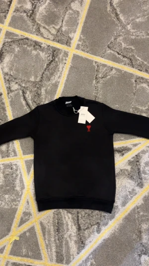 Svart sweatshirt från AMI med rött hjärta - Svart sweatshirt från AMI med broderat rött hjärta och bokstaven A på bröstet. Tröjan har rund hals, lång ärm och diskret AMI-broderi i nacken. Mjuk bomullskvalitet och klassisk passform, perfekt för en clean och stilren look.