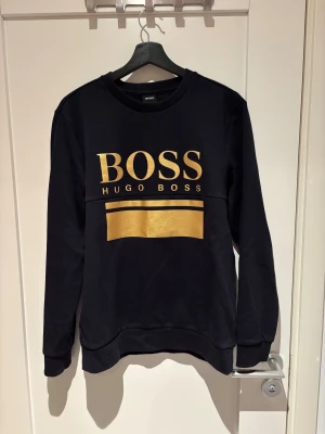 Svart sweatshirt från Hugo Boss med guldtryck - Svart sweatshirt från Hugo Boss med stort BOSS-tryck i guld på bröstet och en bred guldlinje under. Tröjan har knappt används, nästan helt ny. Materialet är mjuk bomull med inslag av polyester för extra komfort.