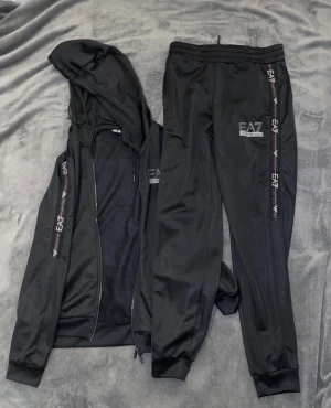 Svarta EA7 mjukisbyxor XS - Svarta mjukisbyxor från Emporio Armani EA7 med logga tryckt på benet och längs sidan. Byxorna har elastisk midja och ribbade muddar vid benslut. Materialet är mjukt och har en lätt glansig finish, perfekt för chill eller träning.