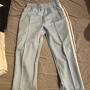 Palm Angels ljusblå mjukisbyxor L - Palm Angels ljusblå mjukisbyxor med vit logga på vänster ben och vita sidoremsor med svart kant. Byxorna har elastisk midja, raka ben och sidofickor. Materialet är en mix av bomull och polyester, vilket gör dem både mjuka och slitstarka.