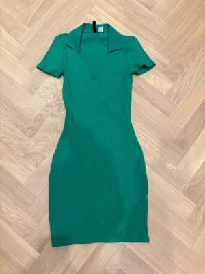 Grön ribbad kortklänning från H&M Divided - Snygg grön ribbad klänning från H&M Divided i storlek XS. Modellen är kort med v-ringning, krage och korta ärmar. Materialet är mjuk bomull med stretch som sitter slim på kroppen. Perfekt för dig som gillar en enkel men trendig look.