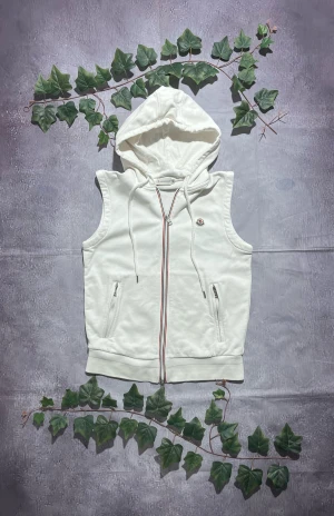 Moncler Gilet i Vit Storlek M [ÄKTA] - 🎨 Färg: Vit 📏 Storlek: Medium (M) ♻️ Skick: 8/10 – vissa mindre bruksspår, men inga större defekter 🧵 Material: Mjukt bomullsblandat sweatshirt-tyg ✨ Detaljer: • Ärm­lös hoodie-väst • Hög krage + huva med dragsko • Två framfickor med dragkedjor • Moncler-patch på bröstet • Dragkedja med röd/vit/blå detalj • Ribbstickad nederkant • Perfekt som lätt ytterplagg eller lager över tröja ‼️ Skickas dagen efter köpet genomförs! ‼️ 