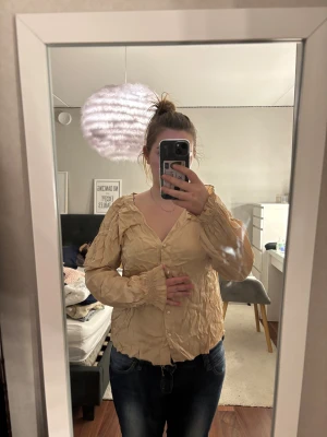 Beige blus med pärlknappar Maja Roos - Supersöt beige blus från Maja Roos med v-ringning och långa puffärmar. Blusen har snygga pärlknappar framtill och ett krinklat tyg som ger cool struktur. Perfekt för dig som gillar unika detaljer och vill sticka ut lite extra.