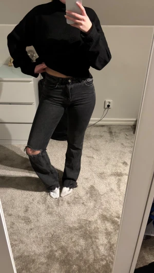 Svarta bootcut jeans med slitning - Säljer ett par svarta jeans med bootcut passform och mid midja. Jeansen har en snygg slitning vid knät och klassiska fem fickor. 
