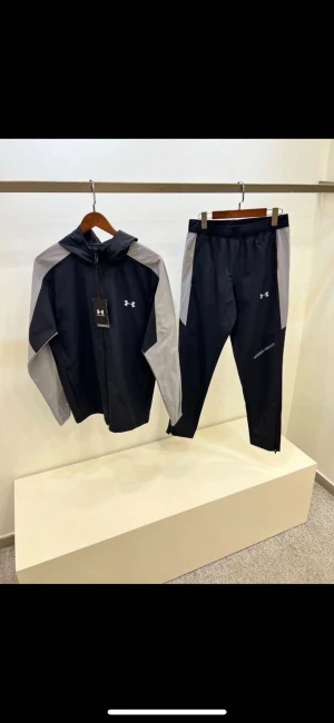 Svarta träningsbyxor från Under Armour - Snygga svarta träningsbyxor från Under Armour med grå paneler längs sidorna. Byxorna har en slim passform och är gjorda i lätt polyester som andas. Perfekta för gymmet eller löprundan, med diskret logga på benet.