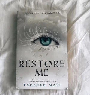Restore Me - En bok med ett mystiskt omslag som visar ett öga omgivet av moln och fjädrar. Perfekt för dig som gillar spännande och känsloladdade berättelser. Slogan på omslaget: 'The past will not forget me'. Skriven av New York Times-bestsäljande författaren Tahereh Mafi.