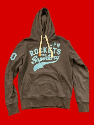 Mörkgrå Superdry hoodie med tryck - Snygg trendig mörkgrå hoodie från Superdry med ljusblå tryck på bröstet och ärmen. Skicka gärna frågor eller funderingar, pris kan ofta diskuteras.
