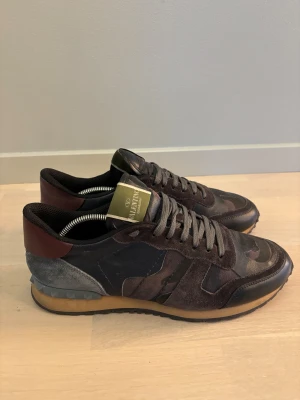 Valentino Garavani Rockrunner sneakers - Snygga Valentino Garavani Rockrunner sneakers i mörka toner med camouflage-mönster. Skorna har detaljer i mocka, skinn och textil, samt en platt sula och rund tå. Grå snörning och coola nitar på hälen ger extra edge. Perfekt för dig som gillar exklusiva streetwear-vibes.