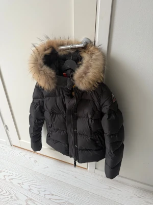 Svart Parajumpers pufferjacka med päls - Svart pufferjacka från Parajumpers med fluffig beige päls runt huvan. Jackan har quiltad design, dragkedja och knappar framtill, samt ribbad detalj i midjan. Insidan har orange detaljer och en praktisk innerficka. Perfekt för kalla dagar.