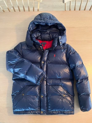 Mörkblå pufferjacka från Polo Ralph Lauren - Snygg mörkblå pufferjacka från Polo Ralph Lauren med röd insida och huva. Jackan har dragkedja och tryckknappar framtill, två stora fickor med knappstängning och ikonisk röd Polo-logga på bröstet. Perfekt för kalla dagar och riktigt chill stil.