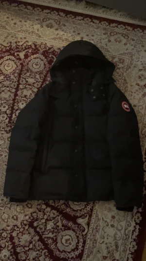 Canada goose wyndham - Säljer en svart dunjacka från Canada Goose med ikonisk röd och vit patch på ärmen. Jackan har huva, dragkedja och knappar framtill, samt quiltad design. Insidan är fodrad med mörkblått tyg och jackan är perfekt för kalla vinterdagar. ÄKTA!!!!