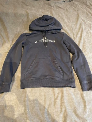 Marinblå hoodie från J.Lindeberg - Snygg marinblå hoodie från J.Lindeberg med vit logga på bröstet. Den är i bra skick och kommer i storlek 150-160.